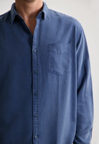 Camasa albastră cu butoni din material de bumbac, cu un buzunar pe pieptul stâng, textură subtilă și mâneci lungi.
