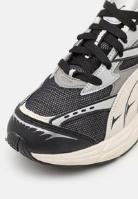 Gros plan sur une sneaker en maille noire et grise avec des lacets noirs, des détails réfléchissants et une semelle blanche avec des accents noirs et gris.