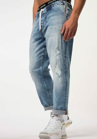 Pantalones jogger de denim desgastado en azul claro con cintura ajustable y detalles de desgarros y decoloración. Combinados con zapatillas blancas y grises.