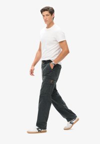 Donkergrijze cargobroek met meerdere zakken, een gele logopatch en een ontspannen pasvorm, gecombineerd met een wit T-shirt en sneakers.