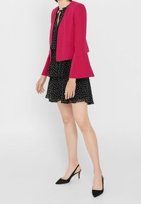 Veste fuchsia à manches cloche portée sur une robe noire à pois. Le mannequin porte des escarpins noirs à bride arrière. Tissu lisse, coupe décontractée, look superposé.