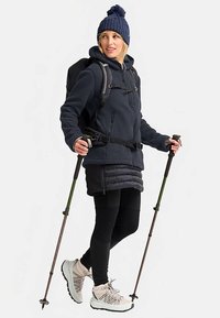 Marineblauwe fleecejas met ritsen, zwarte rugzak, zwarte legging en beige wandelschoenen. Accessoires zijn onder andere wandelstokken en een gebreide muts.