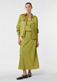 Sukienka z satynowym, oliwkowym zielonym materiałem oraz pasującą spódnicą, z guzikami z przodu i dwiema kieszeniami na piersi, w zestawieniu z czarnymi płaskimi butami.