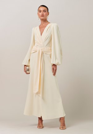 Vrouw in een lange crèmekleurige jurk met opgepofte mouwen, een strikceintuur om de taille, met een kleine gouden clutch in haar hand, en goudkleurige hakken en druppelvormige oorbellen.