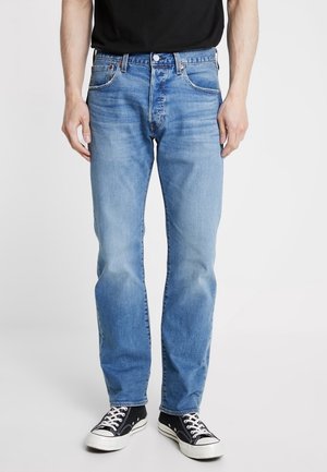 Man wearing light blue straight-leg jeans, black t-shirt, and black low-top sneakers with white laces, standing against a plain background.