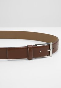 Bruine leren riem met een gladde textuur, rechthoekige zilveren gesp en meerdere gaatjes voor aanpassing. Bevat een subtiele logodetail.