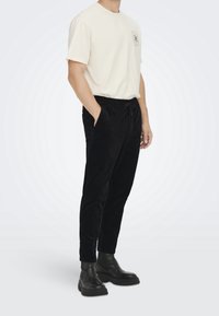 T-shirt en coton beige avec un logo circulaire sur la poitrine, associé à un pantalon en velours côtelé noir et des bottines noires.