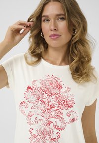 T-shirt crème à coupe décontractée avec des manches courtes, présentant un motif floral rouge vibrant dans un design complexe sur le devant.