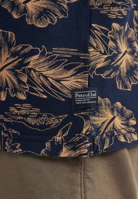 Marinblått tyg med guldblommönster, med en liten "Petrol Ind" etikett och en rak fålldesign. Texturerad yta, avslappnad stil.