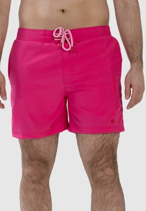 Mann trägt knallpinke Badeshorts mit gebundener Kordel am Bund, steht vor einem einfarbigen hellen Hintergrund.