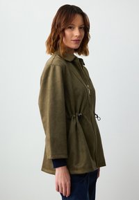 Giacca verde oliva con zip, realizzata in tessuto morbido e testurizzato, con vita cintata, maniche lunghe e colletto a punta. Presenta dettagli con cordino.