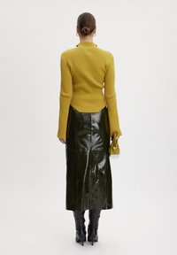 Kobieta z upiętymi włosami nosząca musztardowy sweter w prążki, błyszczącą ciemnozieloną spódnicę midi, czarne botki na obcasie i trzymająca dopasowaną musztardową torebkę.