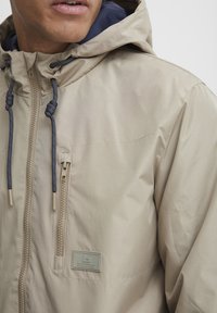 Beige hettejakke laget av glat, vanntett materiale. Har en glidelåslomme og justerbare snorer på hetten.