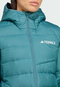 Tirkizna pufasta jakna s prednjim zatvaračem, kvadratnom teksturom i visokim ovratnikom. Sadrži bijeli logotip "TERREX" na lijevoj strani prsa.