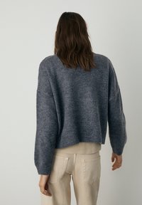 Pull gris raccourci en tissu tricoté doux et texturé avec des manches larges et un ourlet côtelé, associé à un pantalon beige.