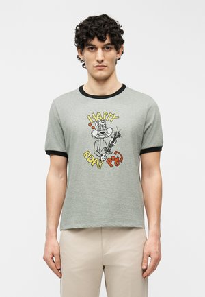 Homme aux cheveux bouclés portant un t-shirt gris avec une bordure noire et un dessin de chaton jouant de la guitare avec le texte « Joyeux anniversaire PSG ».