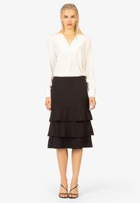 Blusa bianca a maniche lunghe con scollo a V, abbinata a una gonna nera a strati con tre balze e silhouette piatte. Sandali neri con cinturini.