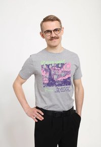 Joven con gafas y bigote que lleva una camiseta gris con un estampado colorido de La Noche Estrellada y una cita, de pie con una mano en la cadera.