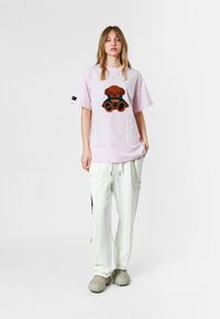 Camiseta oversized rosa clara con un gráfico de oso de peluche, combinada con pantalones de chándal blancos y calzado gris texturizado. Atuendo casual.