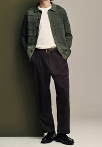 Cardigan verde lavorato a maglia con un motivo testurizzato, abbinato a una maglietta bianca a girocollo e pantaloni larghi scuri, completato da scarpe nere.