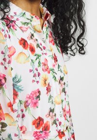 Blumenbluse mit weißem Hintergrund und bunten Blumen in Rosa-, Rot-, Orange- und Grüntönen; kragenförmiges Design aus leichtem Stoff.