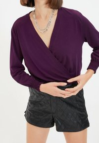 Purple wrap blouse with long sleeves and smocked cuffs, paired with black quilted shorts; smooth fabric, V-neck design, and chain necklace.