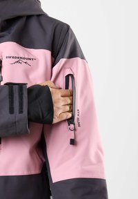 Wasserdichte Jacke mit einem Farbblockdesign in Schwarz und Rosa, ausgestattet mit einer Reißverschlusstasche am Arm und Markenlogo-Details.