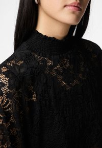 Blusa de renda preta com padrões florais, gola alta franzida e textura semi-transparente, realçando detalhes intrincados e artesanato.