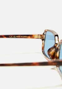Lunettes de soleil en écaille de tortue avec des verres bleus, monture en acrylique transparent, accents en métal doré et marquage "CE UK CA MADE IN CHINA" sur le côté.