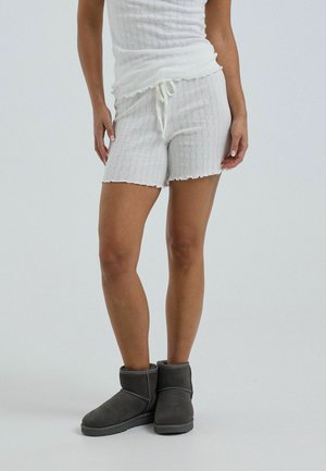 Mujer con pantalones cortos y top de punto blanco con bordes ondulados y botines de color gris oscuro, de pie frente a un fondo liso.
