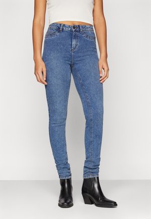 Skinny jeans in medium blue denim, high-waisted design with five pockets, and contrasting stitching. Paired with black ankle boots.