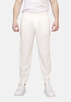 Uomo con pantaloni bianchi a sigaretta con pieghe frontali e sneakers bianche, in piedi su uno sfondo chiaro uniforme.
