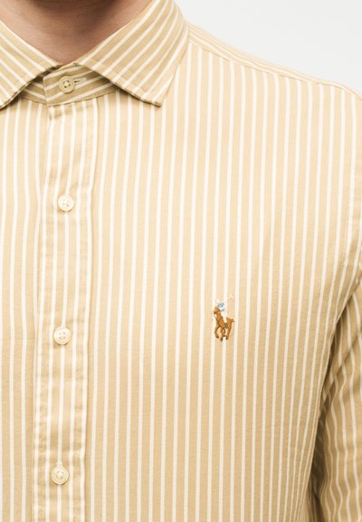 Μπεζ και λευκό πουκάμισο με κάθετες ρίγες και κουμπιά, με ένα μικρό κεντητό λογότυπο παίκτη πόλο στο αριστερό στήθος.