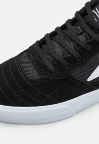 Black suede sneaker featuring mesh accents, textured stitching, and a white rubber sole with a diamond pattern. Flat black laces complete the design.