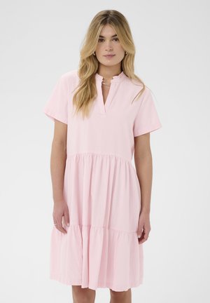 Giovane donna con lunghi capelli biondi che indossa un vestito corto a balze rosa chiaro con collo arricciato, in piedi davanti a uno sfondo semplice.