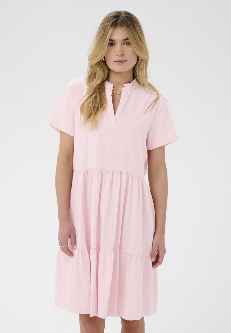 Jeune femme aux longs cheveux blonds portant une robe rose clair à manches courtes, à volants, avec un col à volants, debout sur un fond uni.