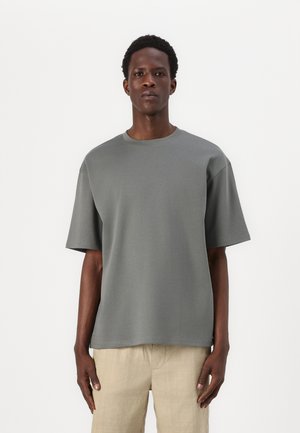 T-shirt gris à manches courtes en tissu doux et lisse, avec un col rond et une coupe décontractée. Aucun motif ni accent visible. Design simple.