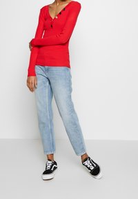 Haut rouge à manches longues côtelé avec un décolleté en V et des boutons noirs, associé à un jean bleu clair droit et des baskets noires.