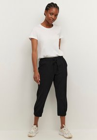 Schwarze, kurze Hosen mit elastischem Bund und Kordelzug, kombiniert mit einem weißen T-Shirt mit kurzen Ärmeln. Die Schuhe sind klobige weiße Sneaker.