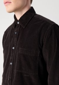 Brown corduroy shirt with a button-down collar, front pocket, and branded logo on the pocket. Features subtle ribbed texture and blue buttons.