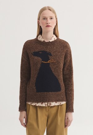 Jeune femme portant un pull marron avec une grande silhouette de chien noir et un collier doré, superposé à une chemise crème à motifs et un pantalon moutarde.