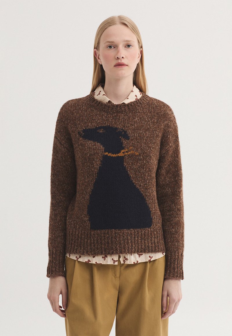 Jeune femme portant un pull marron avec une grande silhouette de chien noir et un collier doré, superposé à une chemise crème à motifs et un pantalon moutarde.