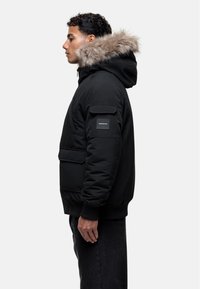Giubbotto piumino nero con cappuccio foderato in pelliccia sintetica, tasca frontale e polsini a costine. Realizzato in un materiale morbido e strutturato con una vestibilità moderna.