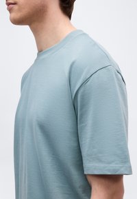 Πλάγια όψη ενός ατόμου που φοράει απλό ανοιχτό μπλε κοντομάνικο πουκάμισο με στρογγυλή λαιμόκοψη σε λευκό φόντο.