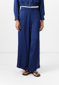 Pantalons larges bleu marine avec une taille élastique et des poches latérales, associés à des tongs noires. Le tissu semble lisse avec une texture douce.