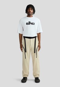 Biała koszula z krótkim rękawem z czarnym logo, zestawiona z beżowymi, puszystymi spodniami z czarnymi paskami i detalami po bokach. Całość dopełniają czarne sneakersy.