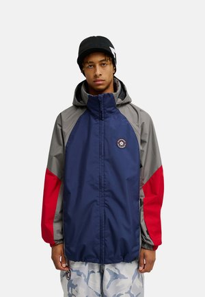 Junger Mann trägt eine farblich abgesetzte Jacke in Blau, Grau und Rot mit Kapuze und eine schwarze Mütze mit weißem Muster.