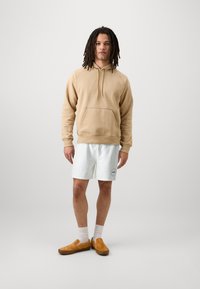Sudadera beige con bolsillo frontal, pantalones cortos blancos con un patrón texturizado y zapatos marrones sin cordones. El modelo está de pie contra un fondo liso.