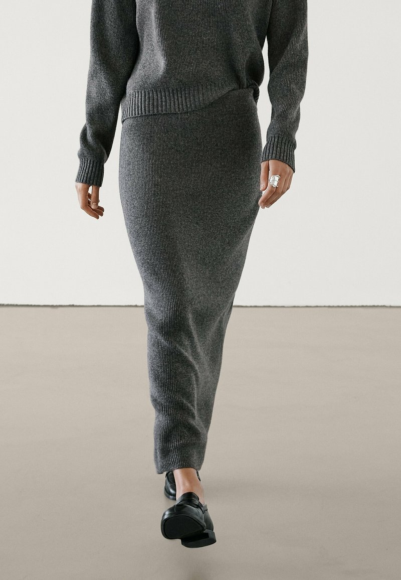 Personne portant un pull en maille gris foncé et une longue jupe assortie, marchant vers l'avant, avec des mocassins noirs et une bague argentée à la main droite.