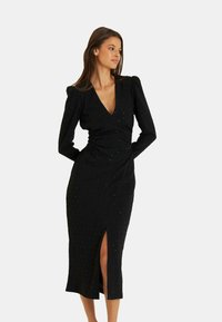 Schwarzes Kleid mit langen Ärmeln, tiefem V-Ausschnitt, gerafften Details, Schlitz auf der linken Seite und verziert mit kleinen, schimmernden Akzenten.